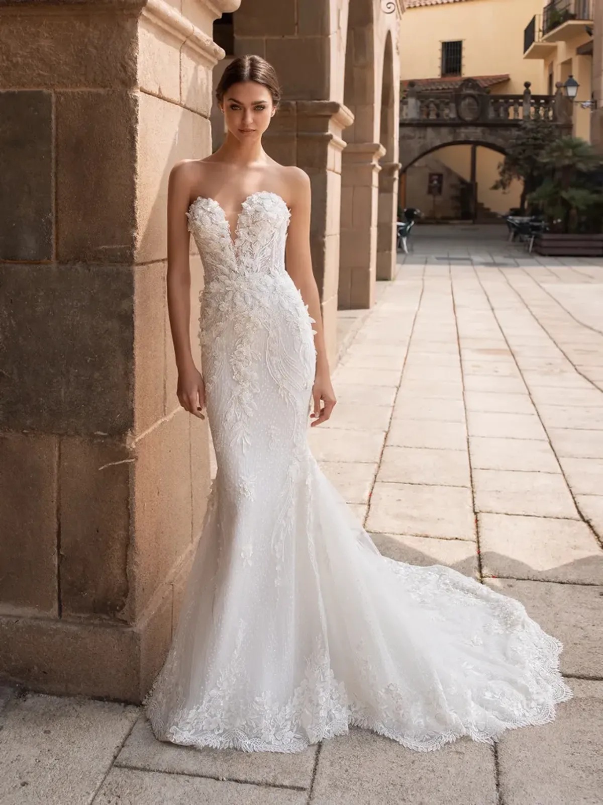 A model showcases the Pronovias Aethra dress near a stone wall in a European old-world cityscape