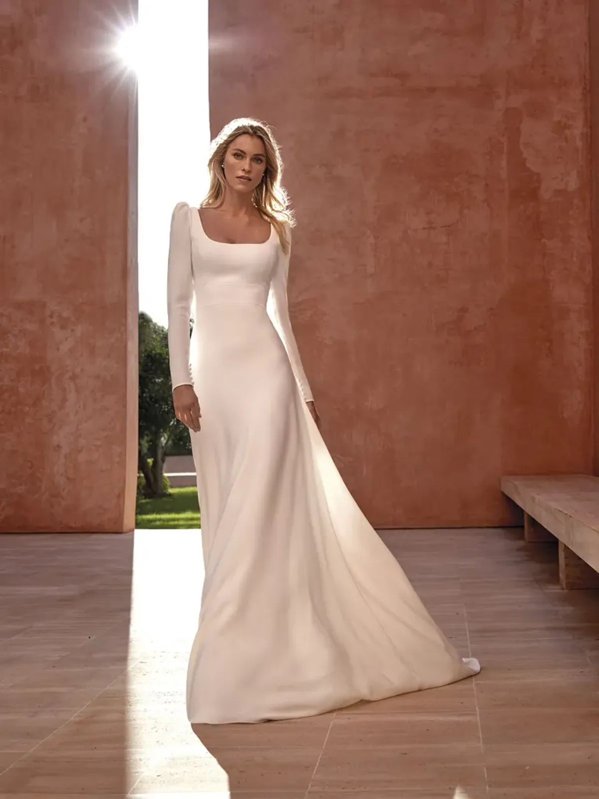 A model showcases the Pronovias Aspen dress in a large corridor with red-brown succo walls and very high ceiling