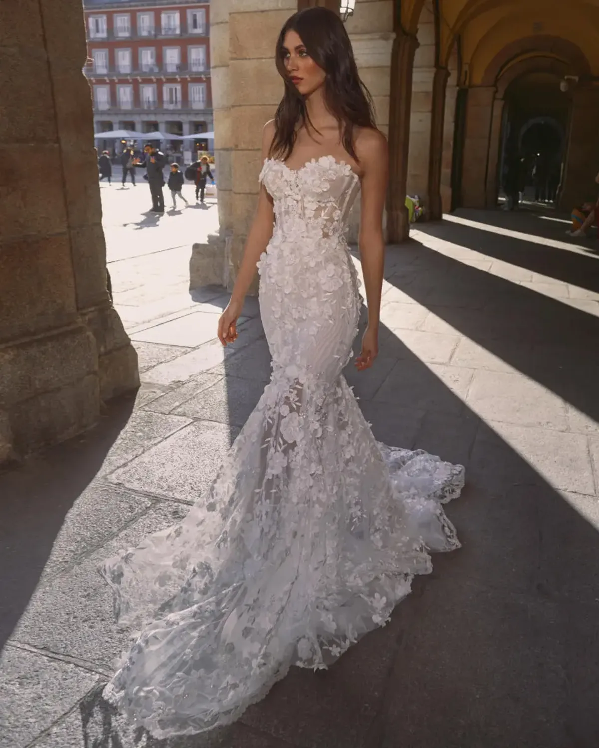 A model showcases the Galia Lahav Carrie dress from the GALA collection in an outside pillared hallway with people in the background