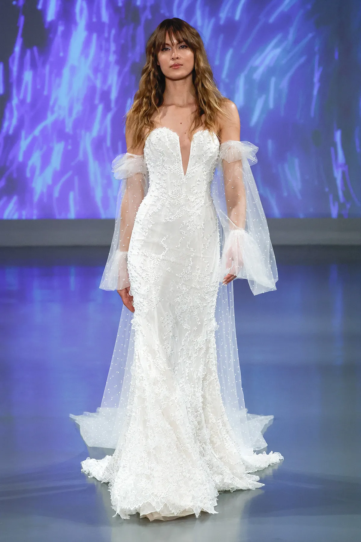 A model showcases the Ines Di Santo Clementine dress while walking on a runway with a blue colored background