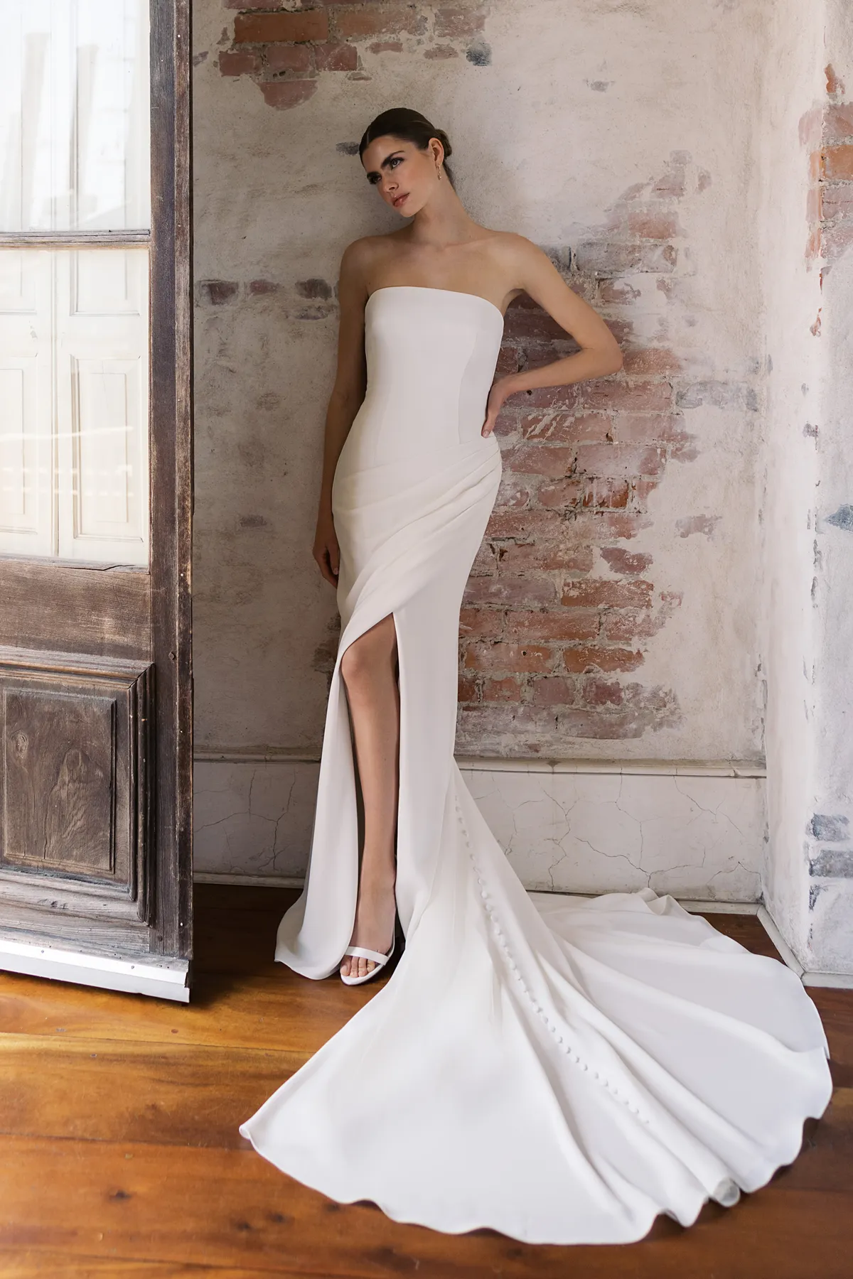 A model showcases the Anne Barge Conti dress from the Blue Willow Bride collection in near an interior wall of a rustic house
