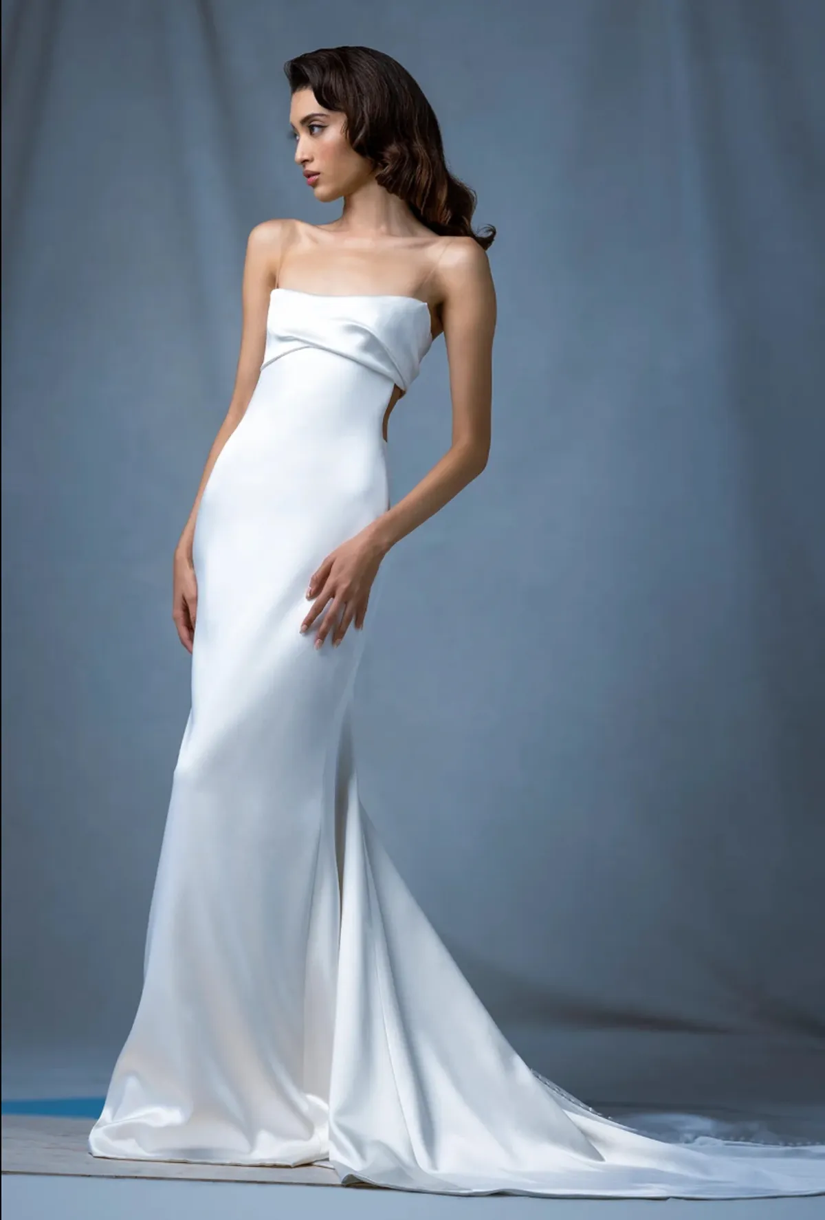A model showcases the Ines Di Santo Dromos dress in a photography studio with a blue-grey backdrop