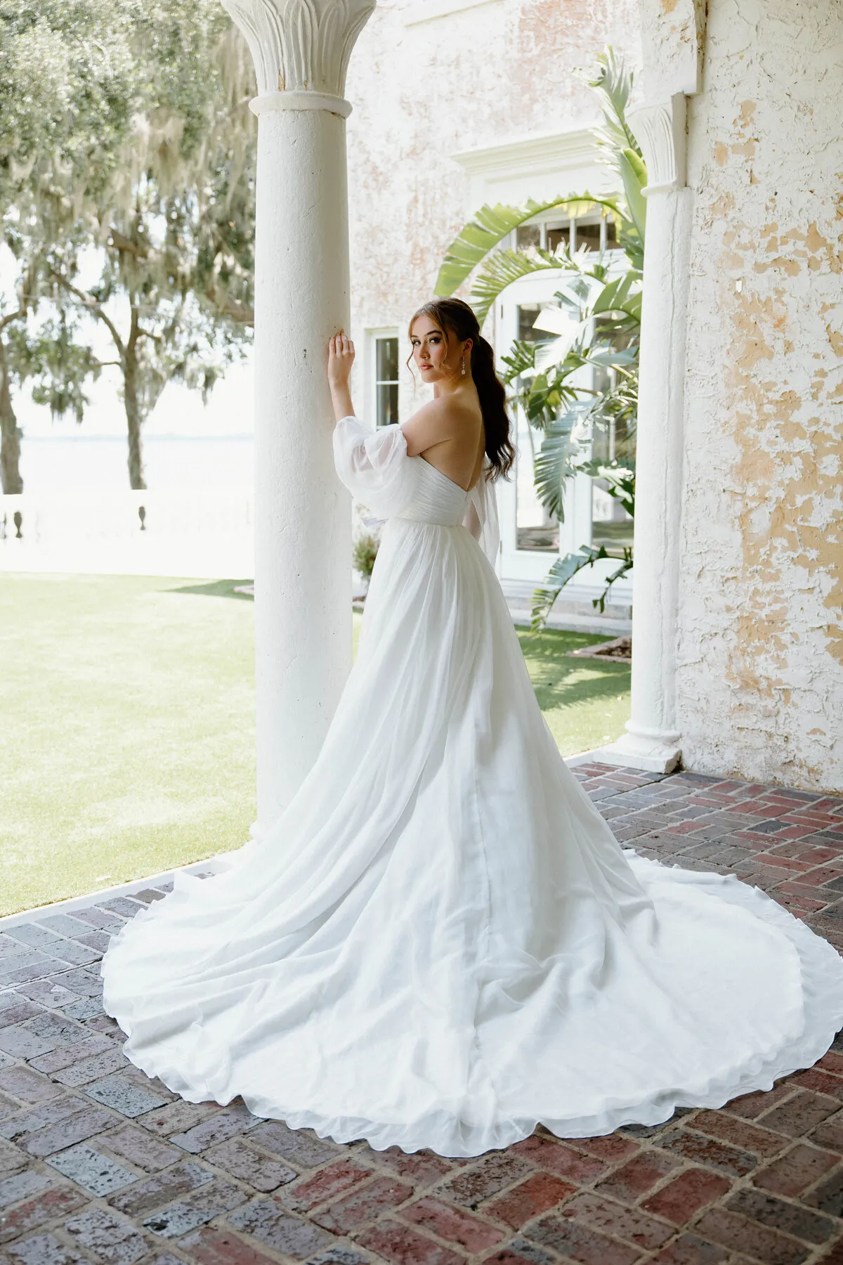 A model showcases the Angel Rivera Freesia dress in front of a stone column outside near a courtyard
