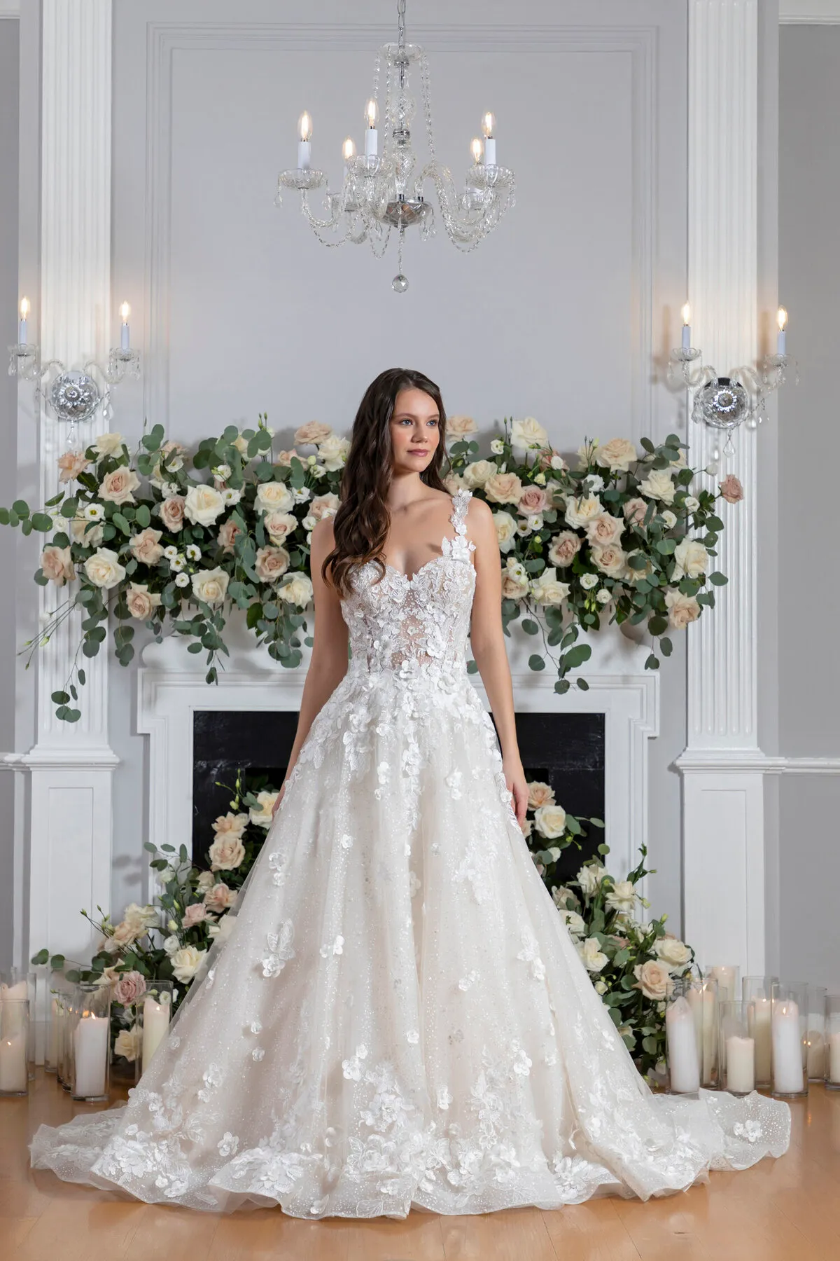 A model showcases the Angel Rivera Garden In Bloom dress in front of a fireplace adorned with flowers and candles