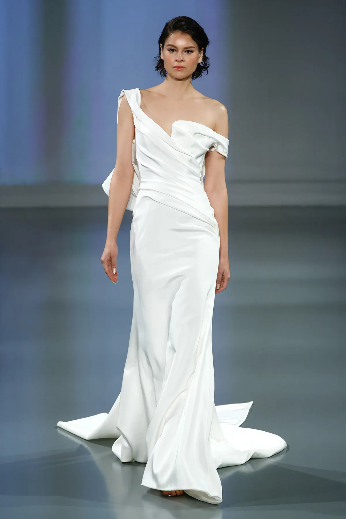 A model showcases the Ines Di Santo Henrietta dress while walking on a runway with a blue colored background
