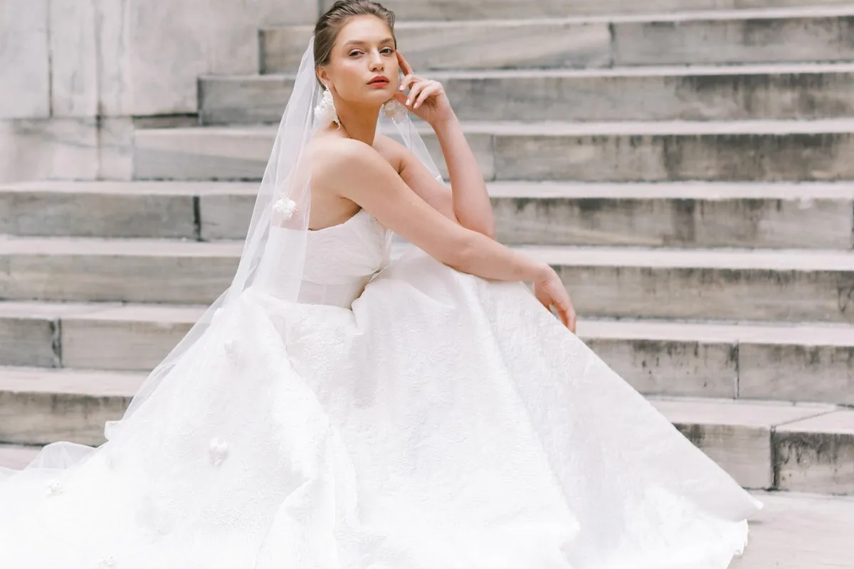 A model sits on a large red velvet chair in a brightly-lit room while wearing an Angel Rivera ballgown
