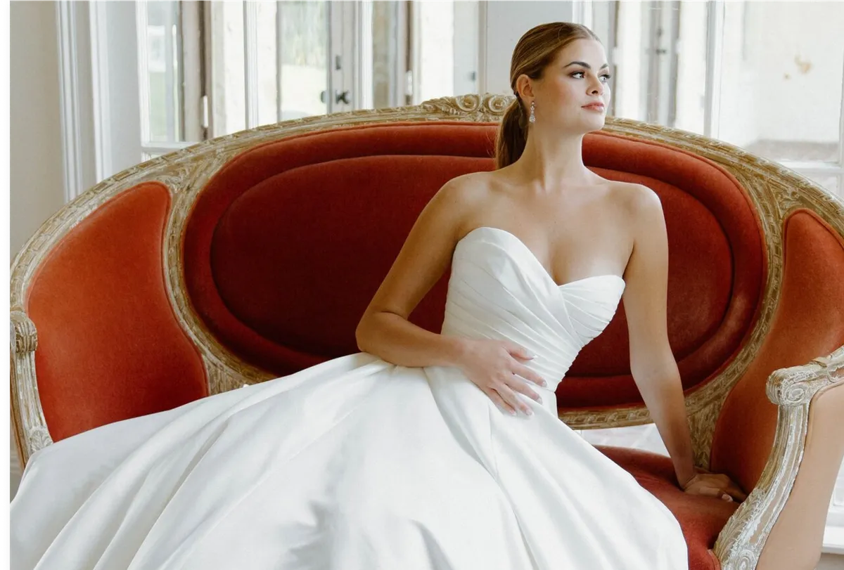 A model sits on a large red velvet chair in a brightly-lit room while wearing an Angel Rivera ballgown