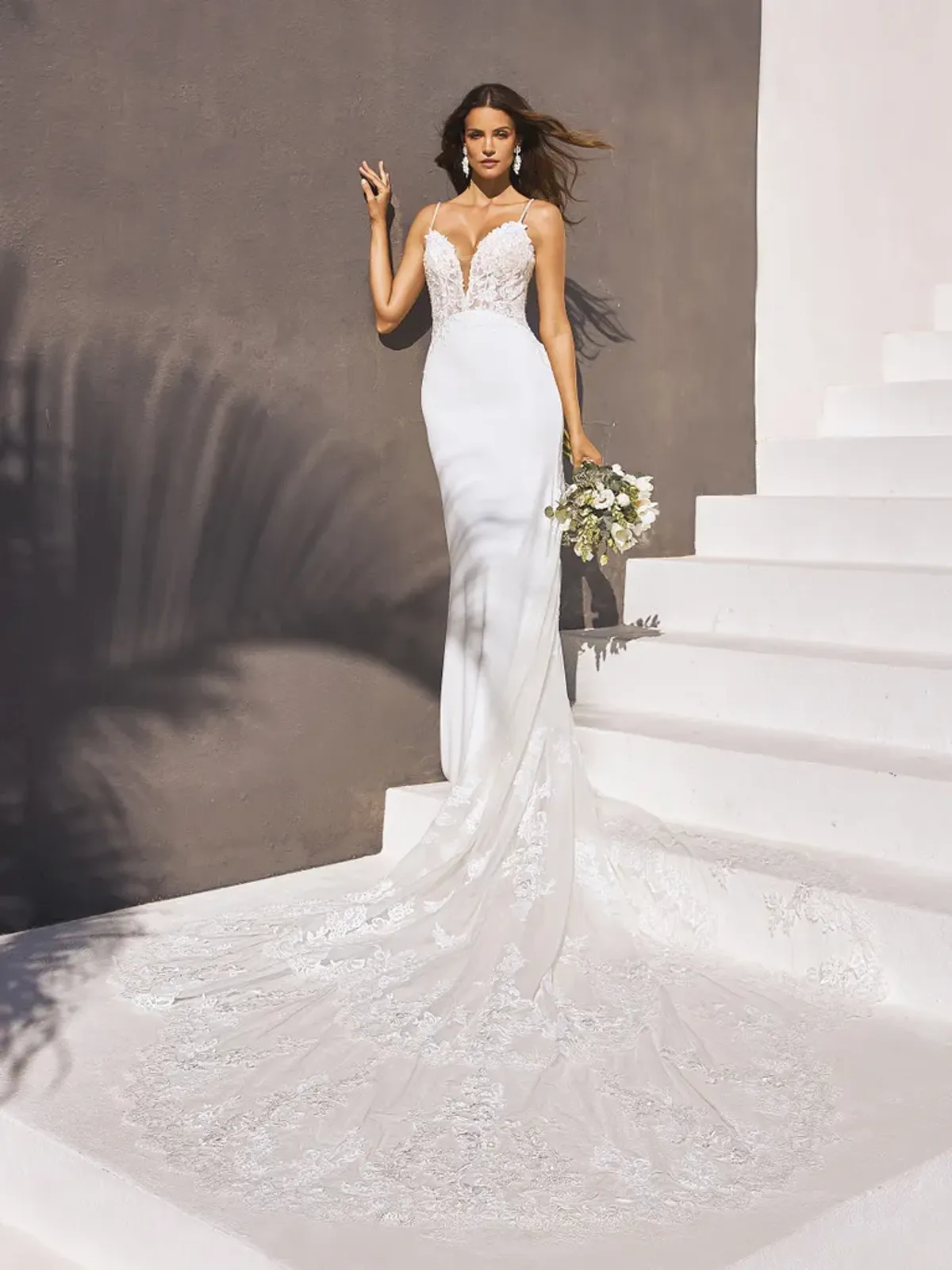 A model showcases the Pronovias Izara dress near a brown wall on white stairs during mid day