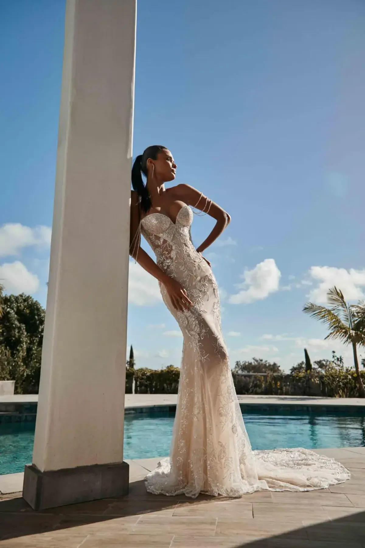 A model showcases the Galia Lahav Nala dress from the Couture collection while leaning on a white column outside near a pool