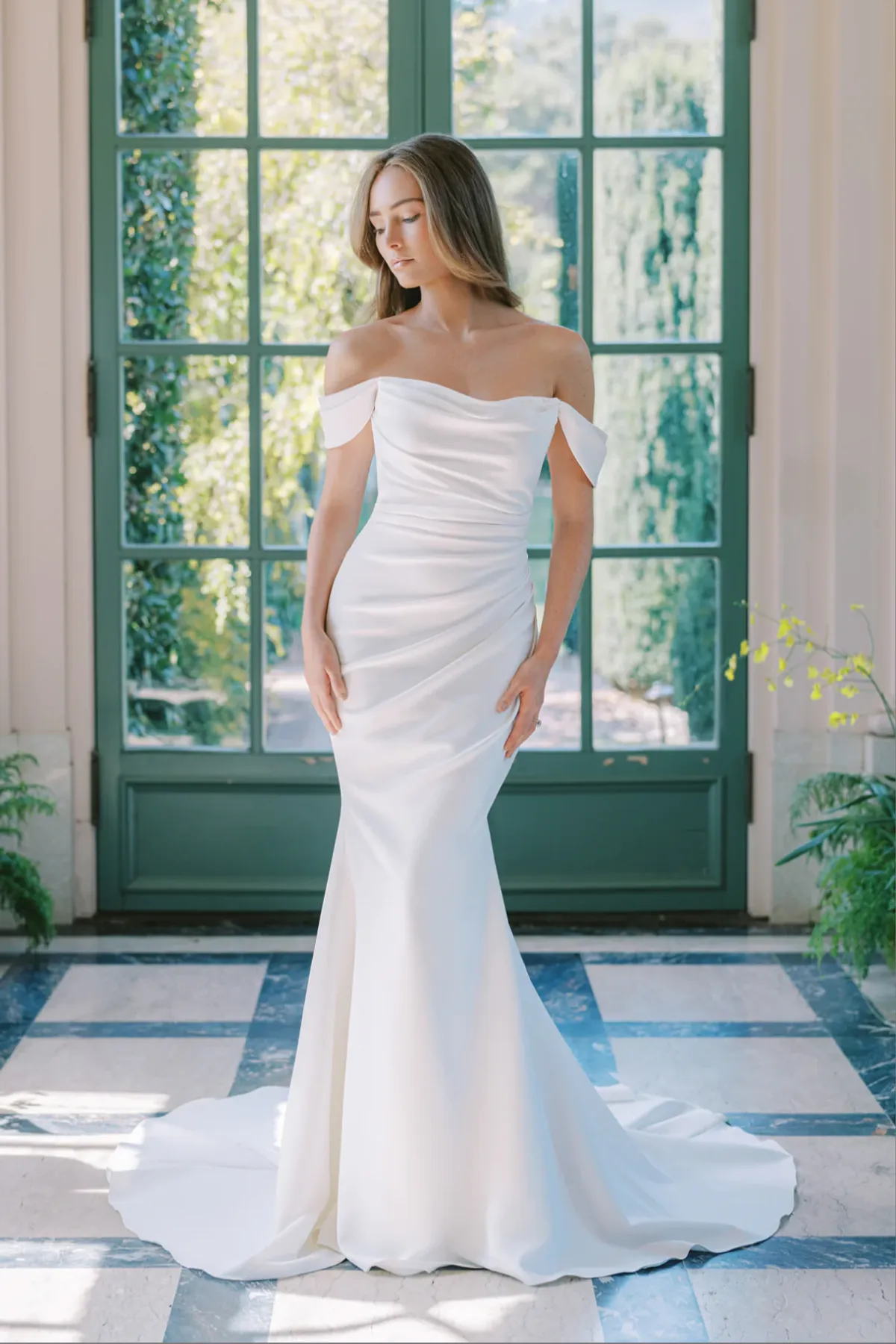 A model showcases the Anne Barge Catalina dress from the Blue Willow Bride collection in front of a large windowed door leading to a garden