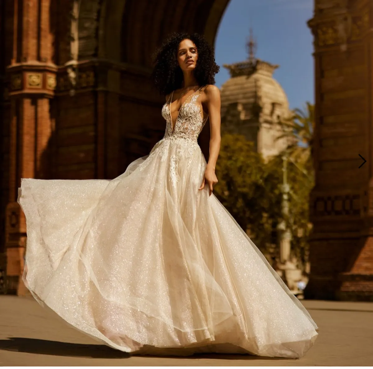 A model showcases the Ines Di Santo Soprano dress from the Ines by Ines Di Santo collection near a large stone arch