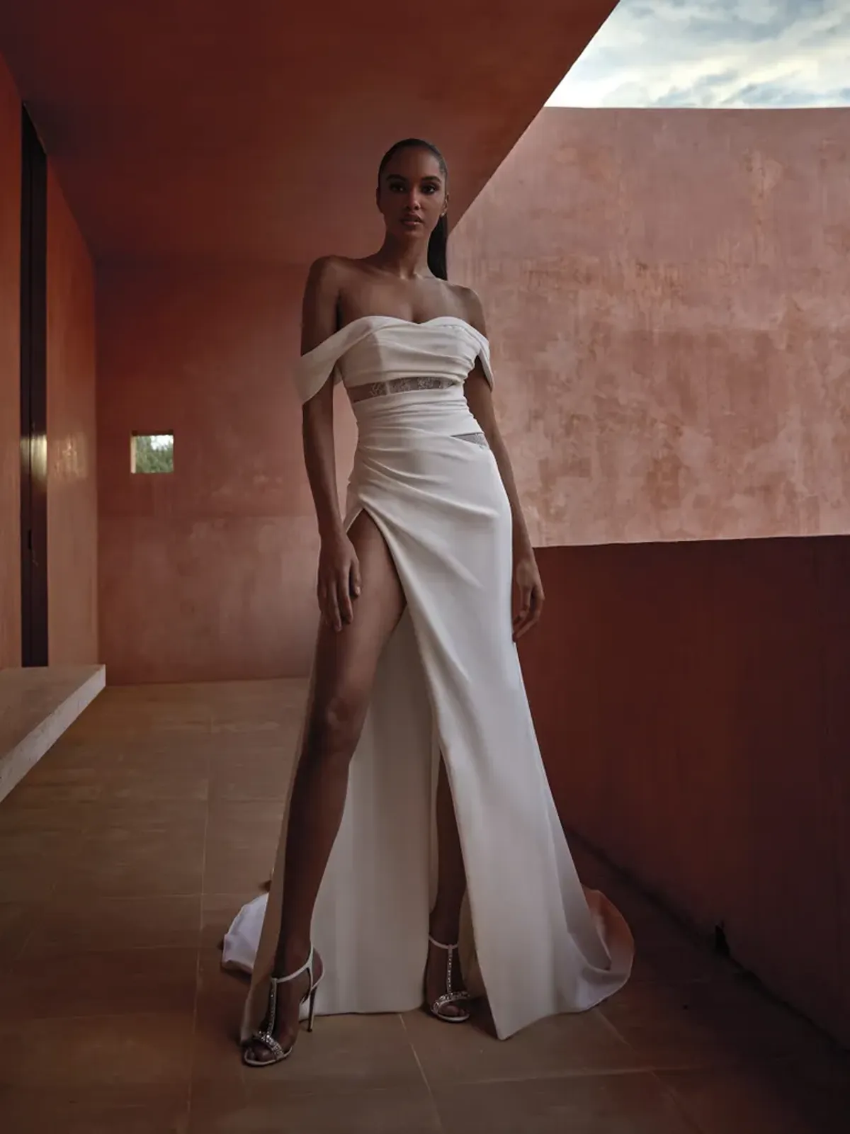 A model showcases the Pronovias Sunj dress on a balcony with red stucco walls