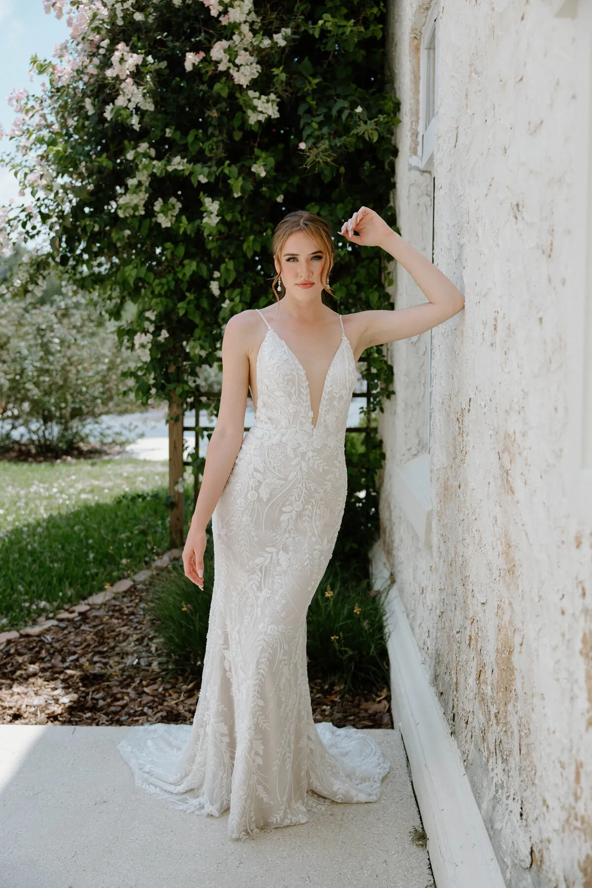 A model showcases the Angel Rivera Willlow dress while leaning on a white stucco wall outside near a flowering tree